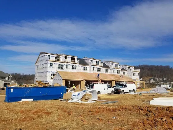 Row of townhouses under construction, wrapped in Tyvek, with exposed framing on the ground floor, construction vans, and a large blue dumpster